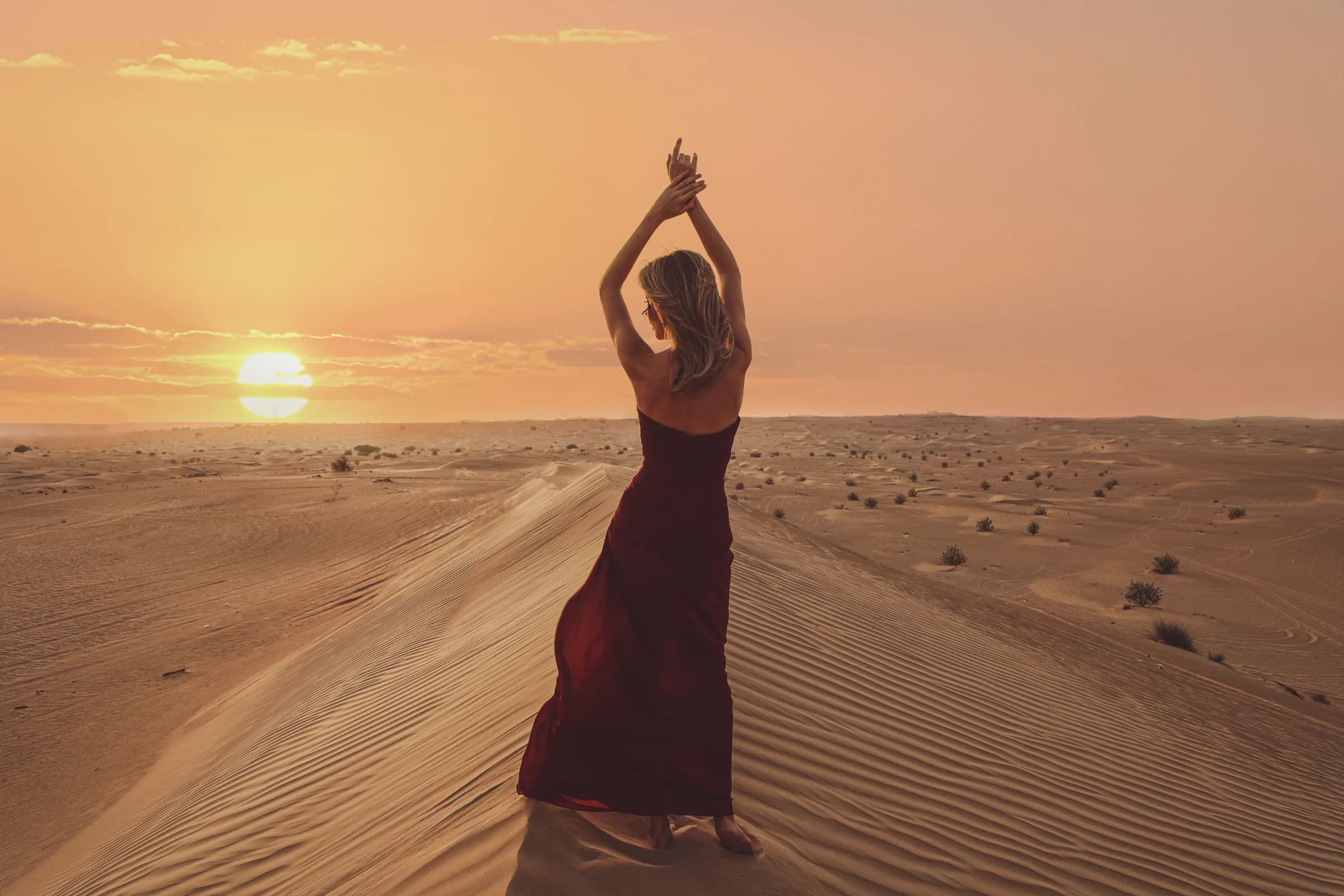 A woman in a red dress poses in the Dubai desert during a stunning sunset with sand dunes stretching into the horizon.